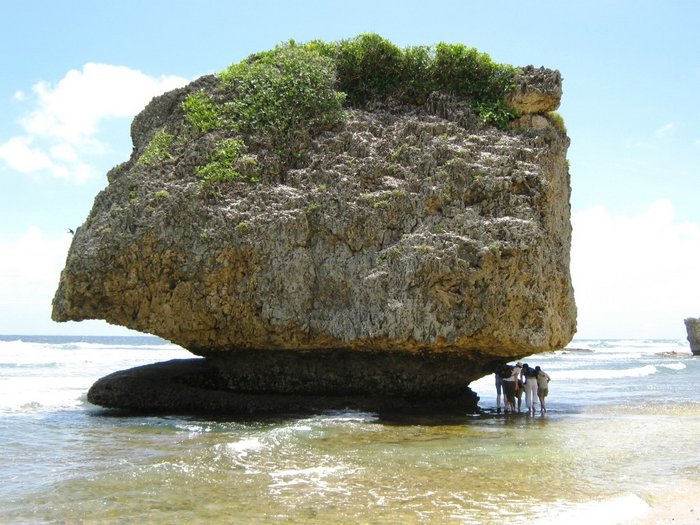 Foto: Gesteinsblock im Meer am Strand. Unregelmäßig geformt und ungefähr zehn Meter hoch. Der unterste, an die Meeresoberfläche grenzende Teil ist eingekerbt. Dort steht eine kleine Gruppe von Menschen und betrachtet den Brocken aus der Nähe.