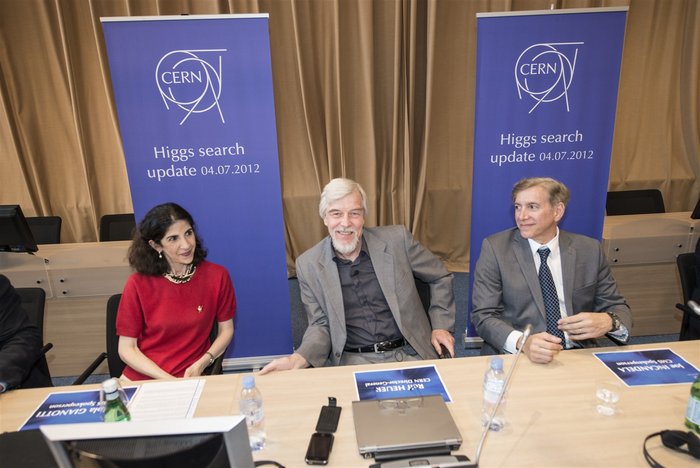 Eine dunkelhaarige Frau, ein grauhaariger Herr und ein blonder Mann sitzen an einem Konferenztisch vor Postern mit der Aufschrift: „Higgs search update 04.07.2012“