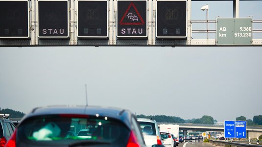 Unten im Bild stehen viele Autos auf einer Autobahn, vom Betrachter aus wegzeigend, hintereinander auf einer Autobahn. Oben ist eine schwarze Tafel, auf der „Stau“ steht und ein rotes Dreieck mit drei Autos abgebildet ist.