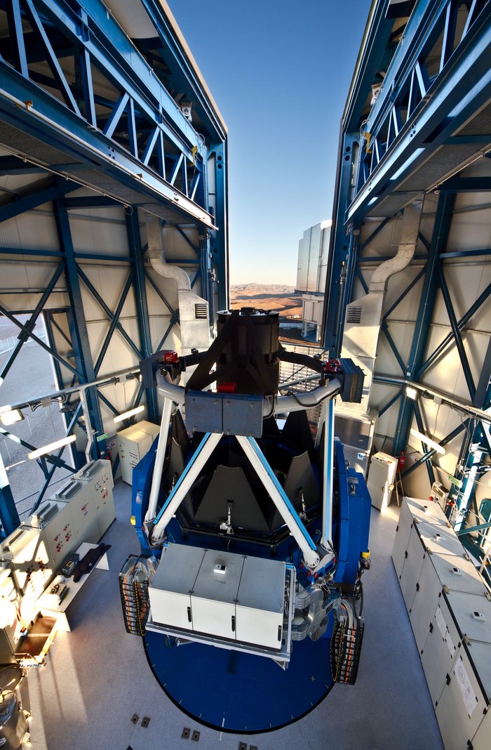 In einem Gebäude mit Metallwänden steht ein zylindrisches Teleskop, senkrecht nach oben gerichtet. Es besteht aus einem tonnenförmigen Unterteil aus Metall, die obere Hälfte besteht aus einem Tubus aus Rohren. An der Seite sind Instrumentenkästen und Flachbandkabel befestigt. Das Gebäude hat in der Mitte einen Spalt, der geöffnet ist. Dahinter ist Wüste, blauer Himmel und ein Teil eines Gebäudes des Very Large Telescopes zu sehen.