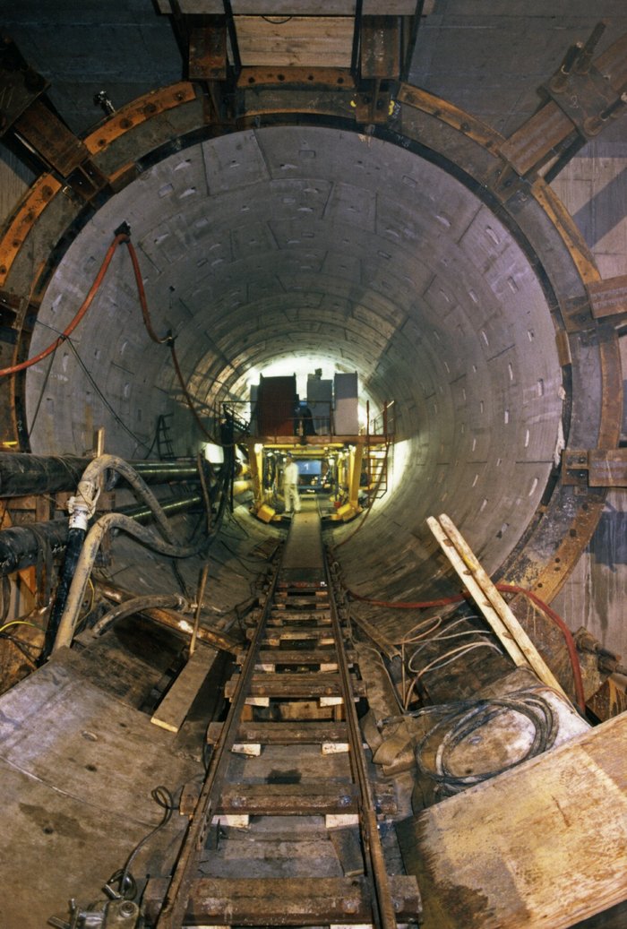 Blick in einen mit Betonelementen verschalten Tunnel, in den ein paar Schienen sowie verschiedene Rohrleitungen und Kabel führen. Im Tunnel ist eine beleuchtete Baumaschine zu erkennen.
