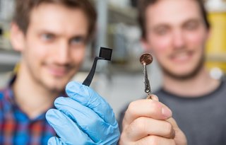 Ein Forscher hält den Mikrochip und ein anderer Forscher ein Ein-Cent-Stück. Beide sind etwa gleich groß.