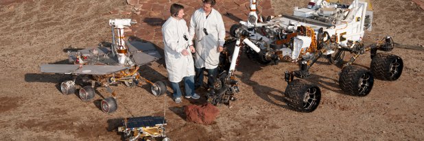 Modelle der drei erfolgreichen Mars-Fahrzeuge auf einem Mars-Testgelände der NASA. Bildquelle: NASA (gemeinfrei) Zwei Männer in weißen Kitteln stehen auf rötlichem, felsig-staubigem Boden. Um sie herum stehen drei Roboter-Fahrzeuge verschiedener Größen, die an Landefahrzeuge für Missionen zu anderen Planeten erinnern.