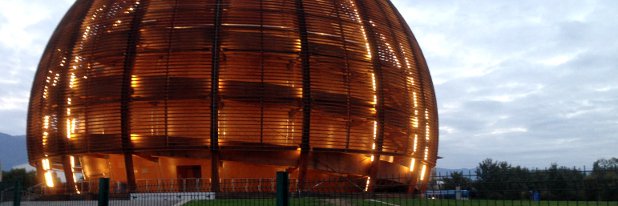 Der Globe of Science and Innovation am Forschungszentrum CERN. Foto: Maria Marinova Ein Gebäude in Form einer unten abgeschnittenen Kugel, umkleidet von Holzlamellen und warm beleuchtet, steht auf einer Wiese.