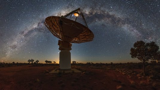 Im Vordergrund befindet sich ein Radioteleskop mit einer großen Schüssel. Im Hintergrund ist die Milchstraße abgebildet.