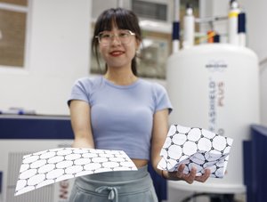 Eine Wissenschaftlerin hält zwei Blätter Papier in der Hand: links ein glattes Blatt mit darauf abgebildeter Atomstruktur, rechts ein leicht zerknüllte Blatt mit darauf abgebildeter Atomstruktur