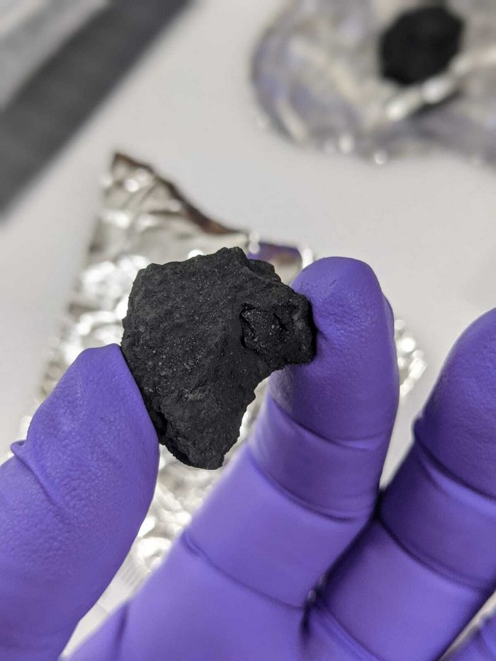 Bruchstück des Winchcombe-Meteoriten Schwarzer Stein zwischen zwei Fingern