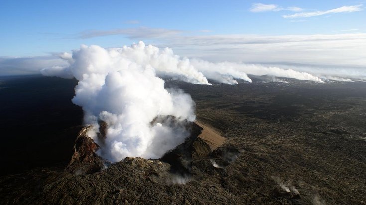 Die Aufnahme zeigt einen Vulkankrater von oben. Aus dem Krater steigt Dampf auf, der weit über das Land zieht.