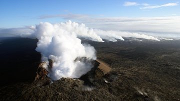 Die Aufnahme zeigt einen Vulkankrater von oben. Aus dem Krater steigt Dampf auf, der weit über das Land zieht.