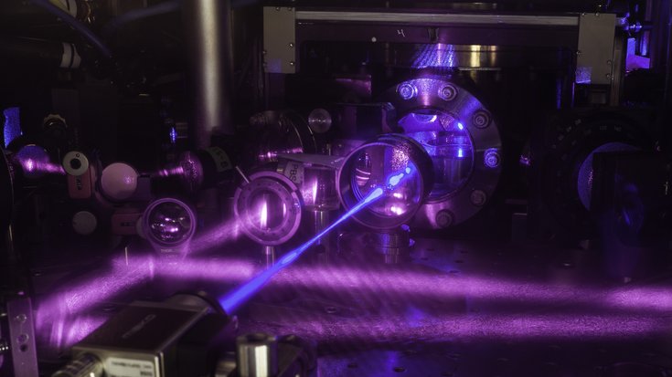 Drei Paare aus Laserstrahlen bilden ein dreidimensionales optisches Gitter, um Strontiumatome für die bisher präziseste Atomuhr der Welt einzufangen, Bildquelle: G. E. Marti/JILA; Lizenz: gemäß den Bedingungen der Quelle Drei violette Laserstrahlen bilden ein optisches Gitter, das von einem blauen Strahl durchzogen wird.