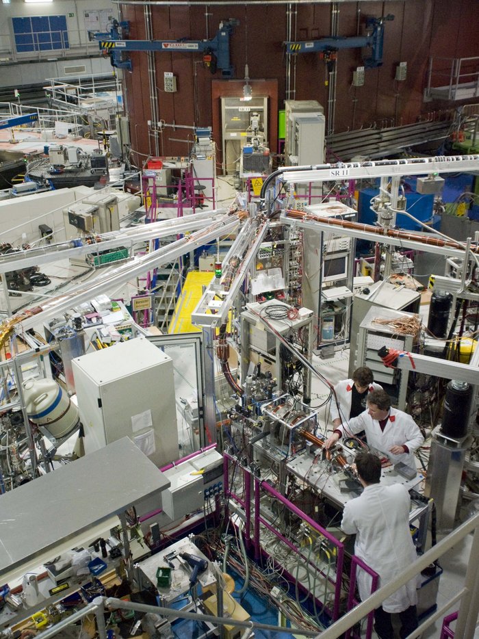 Experimentierhalle um Positronenquelle NEPOMUC Auf dem Bild ist eine große und komplexe Maschine abgebildet, die einen ganzen Raum ausfüllt. Im Vordergrund arbeiten drei Menschen in weißen Kitteln daran.