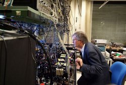 Ein Mann steht vor einer großen Apparatur bei der viele unterschiedliche Kabeln verschiedene elektronische Messgeräte verbinden. Der Mann schaut interessiert in die Apparatur. 