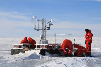Das Bild zeigt fünf Personen in Schneeanzügen, die an einer Forschungsstation arbeiten. 