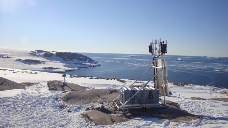 Das Bild zeigt ein GPS Messgerät in der Antarktis. Es ist ein Metallgerüst mit einer grauen Kiste und einigen Geräten zu sehen. Das GPS-Messgerät steht auf einem Felsen. Um den Felsen herum sind Eis und Steine und im Hintergrund ist eine Küste zu seh