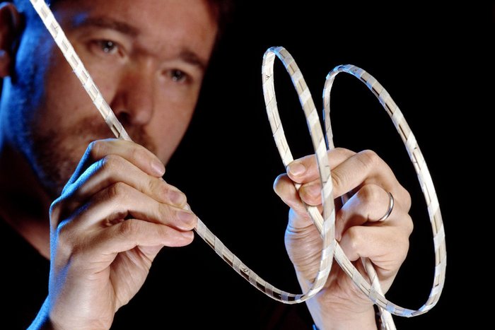 Foto: Forscherkopf vor schwarzem Hintergrund, davor Hände, die ein zentimeterdickes Kabel in mehrere Schlingen legen.