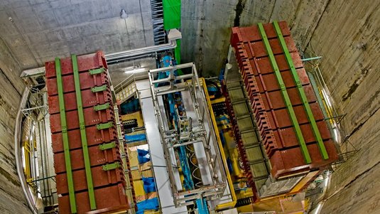 Ansicht eines großen Tunnelschachtes von oben. Unten ist eine große Maschine aus schweren Eisenträgern und mit vielen Kabeln zu sehen, um die herum eine Eisentreppe und Laufwege führen.