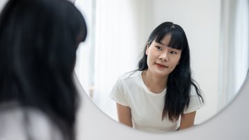 Eine junge Frau blickt in ihr Spiegelbild in einem runden Spiegel. Sie hat langes, glattes, dunkles Haar mit Pony und trägt ein weißes T-Shirt. 