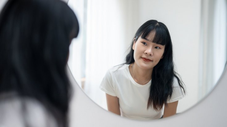 Eine junge Frau blickt in ihr Spiegelbild in einem runden Spiegel. Sie hat langes, glattes, dunkles Haar mit Pony und trägt ein weißes T-Shirt. 