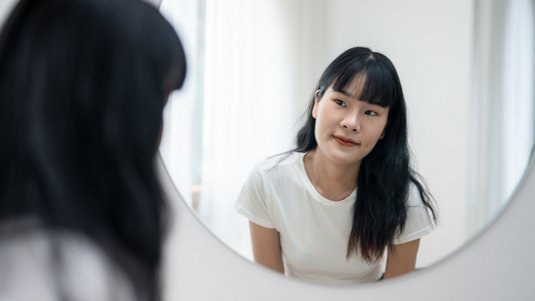 Eine junge Frau blickt in ihr Spiegelbild in einem runden Spiegel. Sie hat langes, glattes, dunkles Haar mit Pony und trägt ein weißes T-Shirt. 
