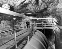 Ein Schwarz-Weiß-Foto zeigt eine unterirdische Höhle, in der einer großer metallischer Tank steht. Über dem Tank führen metallene Laufwege entlang, auf einem davon steht ein Mann in Bergarbeiter-Schutzkleidung.