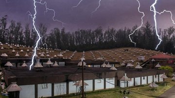 Mehrere flache Gebäude, die eine Art Antenne auf dem Dach haben, stehen in einer dunklen Landschaft. Über ihnen breiten sich Blitze aus.