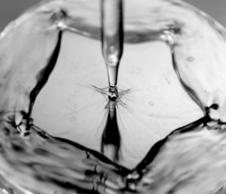 Eine klare Flüssigkeit läuft aus einer Glaspipette auf eine glatte, feste Oberfläche. Auf der Oberfläche bildet sich aus der Flüssigkeit ein Fünfeck.