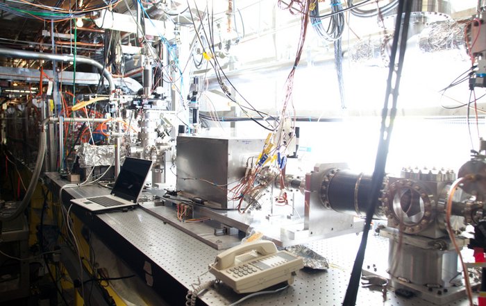 Experiment zur Kielfeld-Beschleunigung am SLAC Eine metallene Kammer steht auf einem Tisch, umgeben von sehr vielen Kabeln und Anschlüssen. Durch die kurzen Seiten der quaderförmigen Kammer läuft ein Rohr aus dem Bild heraus.