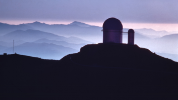 Siluhette einer Teleskopkuppel auf einem Berg, im Hintergrund entferntere Berge und Wolken