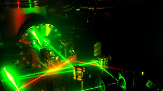 Linsen, Blenden, Spiegel und andere optische Elemente leiten grüne und rot-orange Laserstrahlen entlang eines Experimentier-Tischs.