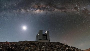 Auf einem steinigem Untergrund steht ein Teleskopgebäude. Es ist Nacht, im Hintergrund scheint der Vollmond und ein deutlich sichtbarer Sternenhimmel breitet sich über dem Bau aus.