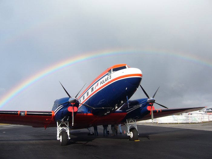 Das zweimotorige Flugzeug ist orange-weiß-blau gestreift. Es steht auf einem Rollfeld. Im Hintergrund wird es von einem Regenbogen umrahmt.