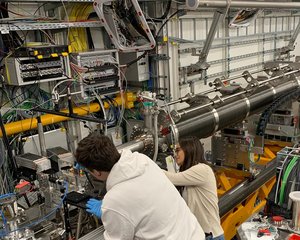 Zwei Forschende arbeiten mit Schutzhandschuhen an einer Maschine, von der eine lange metallene Röhre ausgeht.