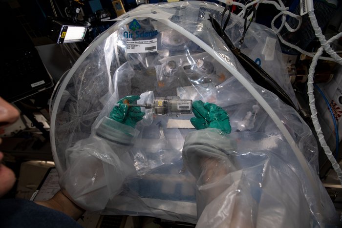 Matthias Maurer mischt Beton auf der ISS In einem Plastikzelt halten zwei behandschuhte Händen einen transparenten Kolben mit einer grauen Masse darin