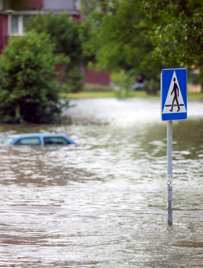 Hochwasserüberflutete Straße Foto. Fluss bei Hochwasser. Im Vordergrund Verkehrszeichen im Wasser stehend, im Hintergrund ein abgesoffenes Auto..