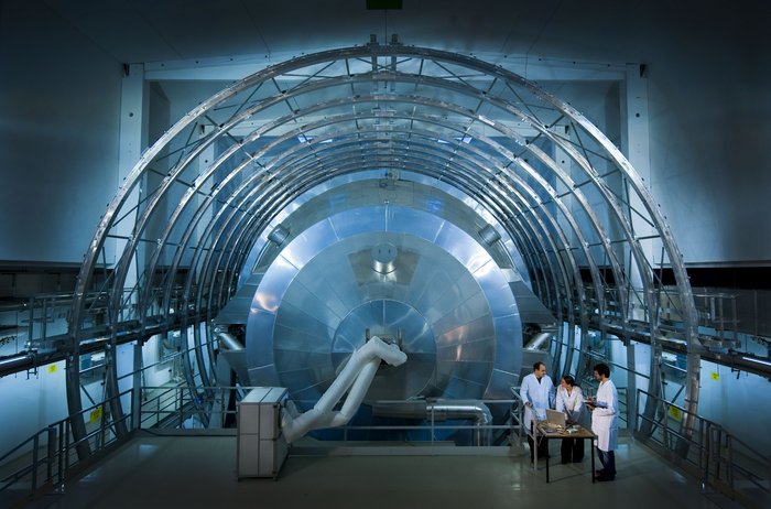 Das KATRIN Experiment Das Spektrometer ist fertig installiert in der Montagehalle. Davor befinden sich drei Wissenschaftler an einem Tisch, auf dem ein Computer steht.