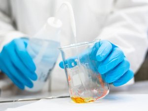 Eine Person hält in einem Labor in einer behandschuhten Hand einen gläsernen Becher, in der eine Flüssigkeit golden scheint
