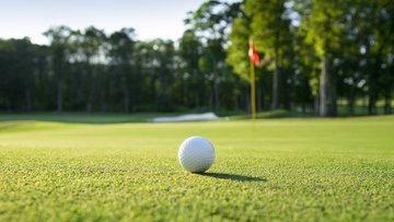 Nahaufnahme eines weißen Golfballs auf saftig grünem Rasen, im Hintergrund unscharf eine rot-weiße Fahne, ein Sandbunker und dichte Bäume unter blauem Himmel.