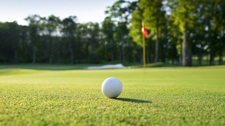 Nahaufnahme eines weißen Golfballs auf saftig grünem Rasen, im Hintergrund unscharf eine rot-weiße Fahne, ein Sandbunker und dichte Bäume unter blauem Himmel.