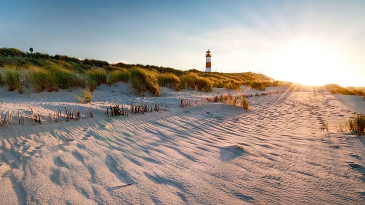 Sandstrand mit Dünen, im Hintergrund ein rot-weiß gestreifter Leuchtturm