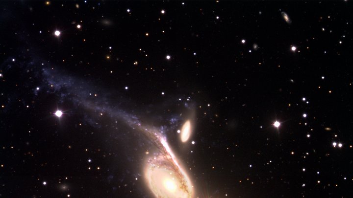 Vor dunklem Hintergrund eine helle Spiralgalaxie mit zwei langgestreckten Armen, rechts des oberen Armes eine kleine ovale Galaxie; rechts unterhalb des Zentrum der Spiralgalaxie leuchtet ein helles, rötliches Objekt