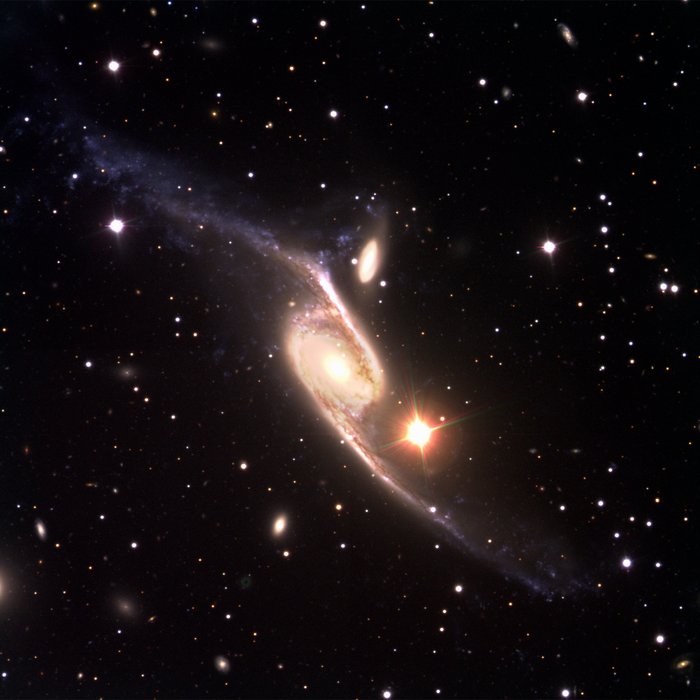 Kosmische Kollision Vor dunklem Hintergrund eine helle Spiralgalaxie mit zwei langgestreckten Armen, rechts des oberen Armes eine kleine ovale Galaxie; rechts unterhalb des Zentrum der Spiralgalaxie leuchtet ein helles, rötliches Objekt.