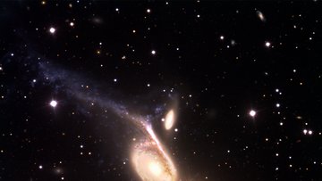 Vor dunklem Hintergrund eine helle Spiralgalaxie mit zwei langgestreckten Armen, rechts des oberen Armes eine kleine ovale Galaxie; rechts unterhalb des Zentrum der Spiralgalaxie leuchtet ein helles, rötliches Objekt