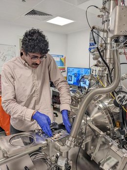 Männliche Person steht vornübergebeugt an einem Tisch, auf dem verschiedene metallene Apparaturen aufgebaut sind. Diese sind mit Kabeln miteinander verbunden. Im Hintergrund steht ein Computer.