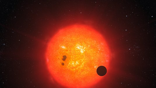Stern mit hellen und dunklen Regionen, davor als schwarzer Kreis ein Planet.