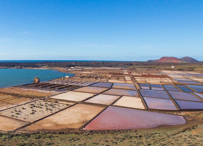 Salzgewinnung auf Lanzarote Foto: An einer Küste sind mit Steinen abgetrennte, quadratische Wasserbecken zu sehen, daneben Haufen weißen Salzes.