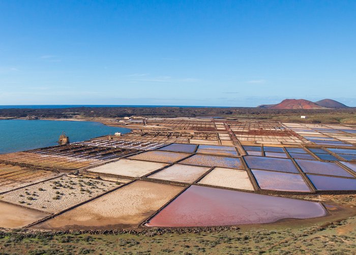 Foto: An einer Küste sind mit Steinen abgetrennte, quadratische Wasserbecken zu sehen, daneben Haufen weißen Salzes.