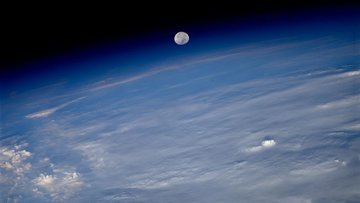 Blick aus dem All auf die bewölkte Erde; die Atmosphäre ist als milchige Schicht erkennbar; am äußeren Rand der Atmosphäre ist der Mond zu sehen