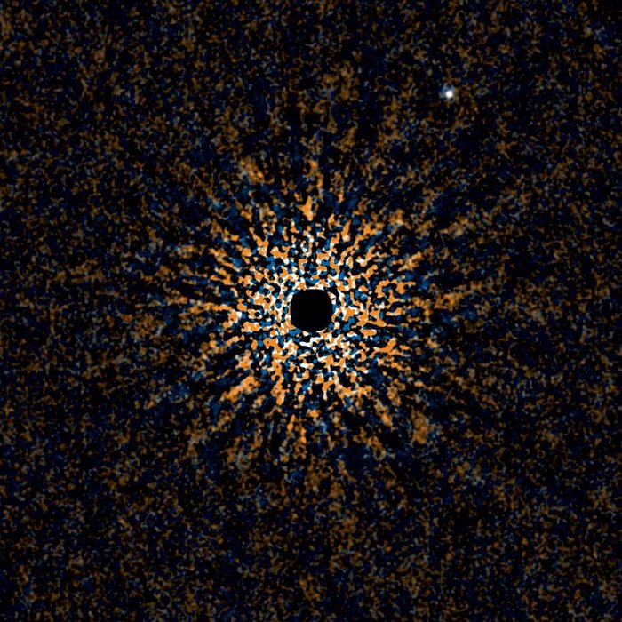Das Bild zeigt in der Mitte einen runden schwarzen Fleck, der strahlenförmig von weißen, orangefarbenen und blauen Flecken umgeben ist. Oben rechts hebt sich ein etwas verschwommener weißer Punkt vom Hintergrund ab.