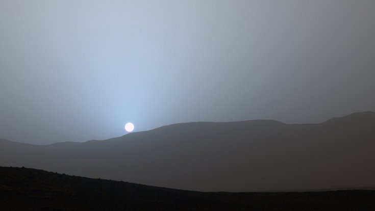 An einem Himmel steht ein hell leuchtender Himmelskörper über einer grauen, bergigen Landschaft
