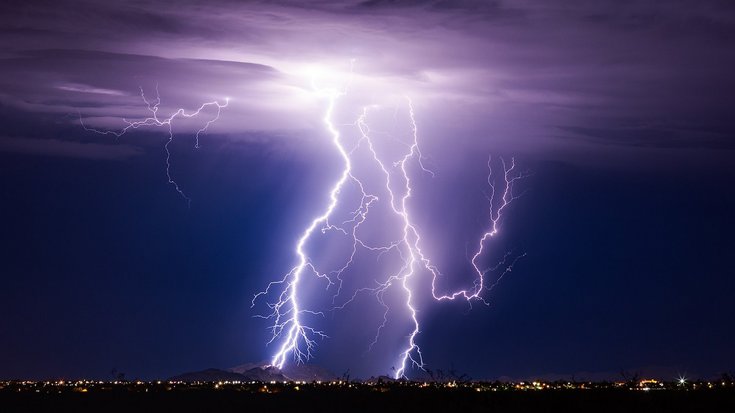 Nachtaufnahme eines Gewitters über einer Stadt: Mehrere verzweigte, hell leuchtende Blitze durchziehen den violett-dunkelblauen Himmel und schlagen in einen Hügel und die Landschaft ein. Am Horizont sind zahlreiche kleine Lichter der Stadt zu erkennen.